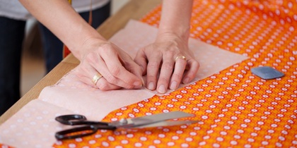 Händer som nålar fast mönsterpapper på orange tyg med sax och krita bredvid.