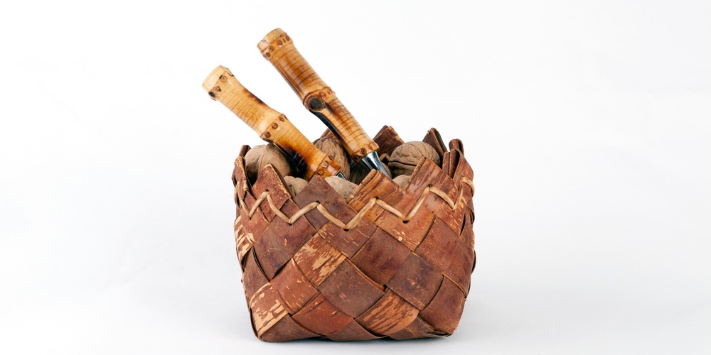 En flätad korg av bark med två knivar med trähandtag i, mot vit bakgrund.