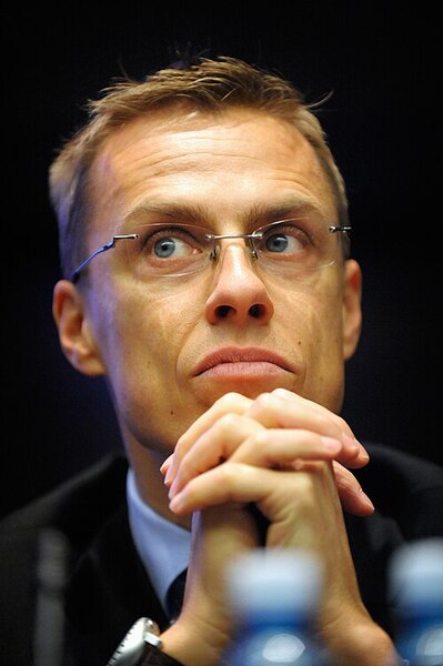 En person i kostym sitter med händerna knäppta framför sig vid ett bord med vattenflaskor.