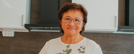 Kvinna står i kök bakom ett bord med olika grönsaker och rotfrukter.