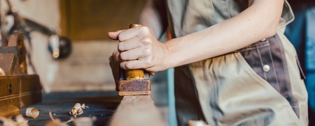 En person i arbetskläder hyvlar trä i en snickarverkstad, fokus på händer och verktyg.