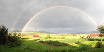 Landskap med gröna fält, bondgårdar och en tydlig dubbel regnbåge mot en molnig himmel.