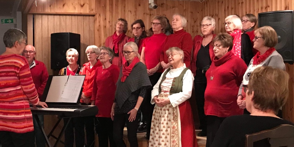 En kör står på en liten scen och sjunger, ledd av en dirigent vid ett elpiano. Publik sitter framför.