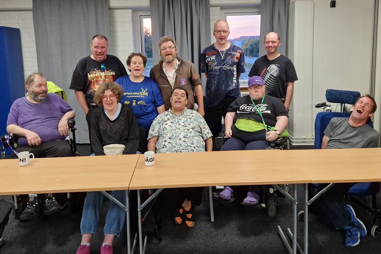 En grupp på tio personer, några i rullstol, sitter och står runt ett bord i ett mötesrum och ler mot kameran.