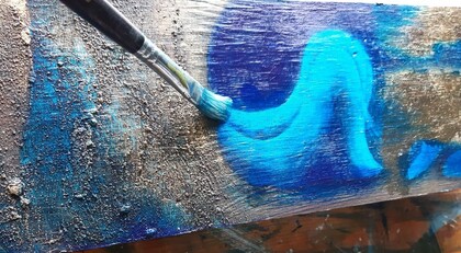 En pensel målar ett blått mönster på en grov träyta med blå och turkosa nyanser.