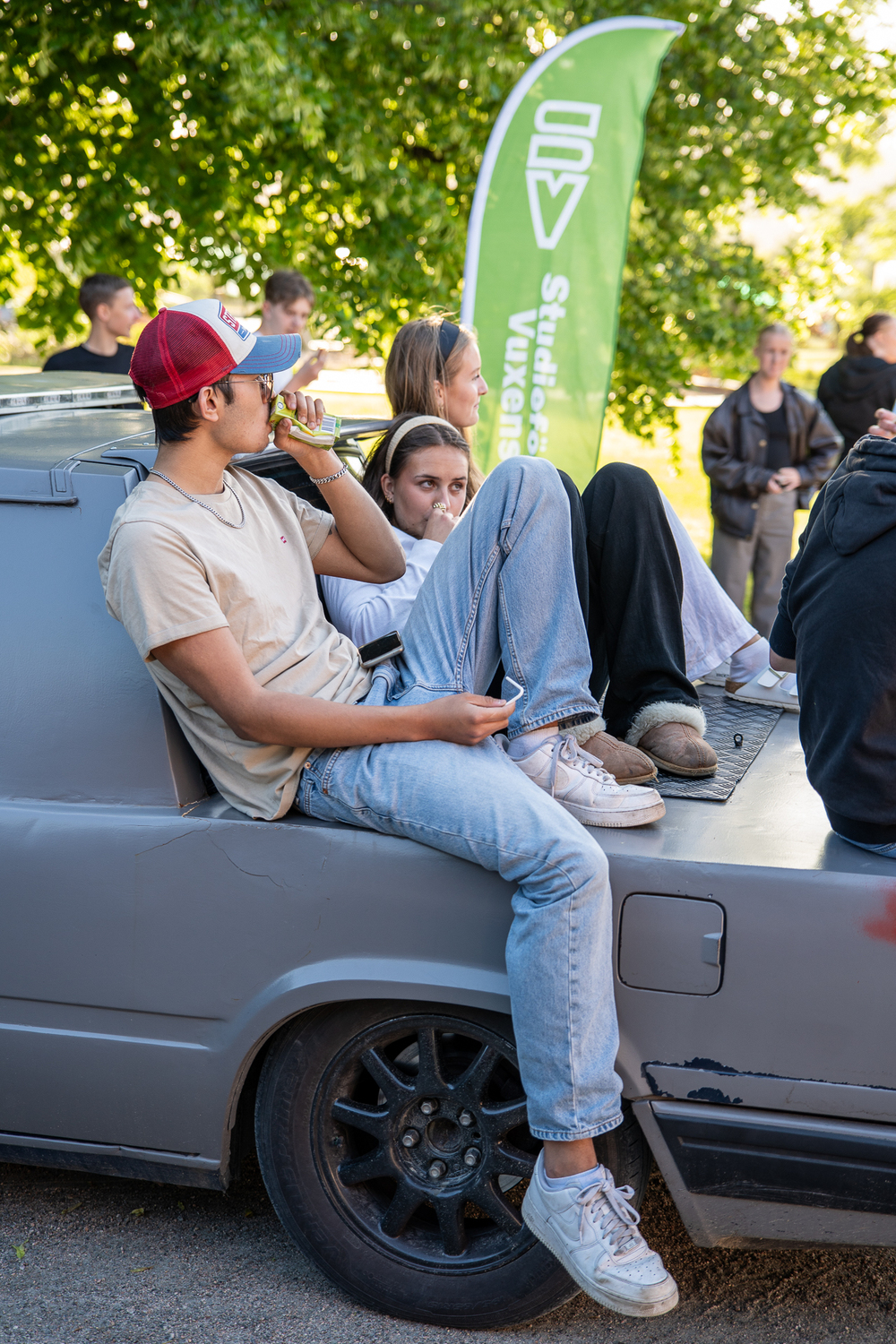 Ungdomar sitter avslappnat på flaket av en grå bil under ett utomhusevenemang.