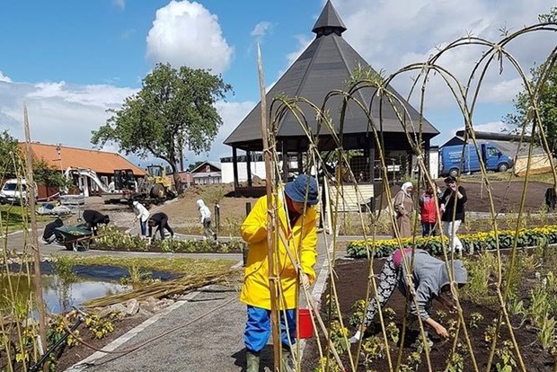Flera personer arbetar i en trädgård nära en damm och ett lusthus under en solig dag.