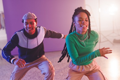 Två unga personer dansar energiskt i ett färgglatt rum med lila bakgrund.