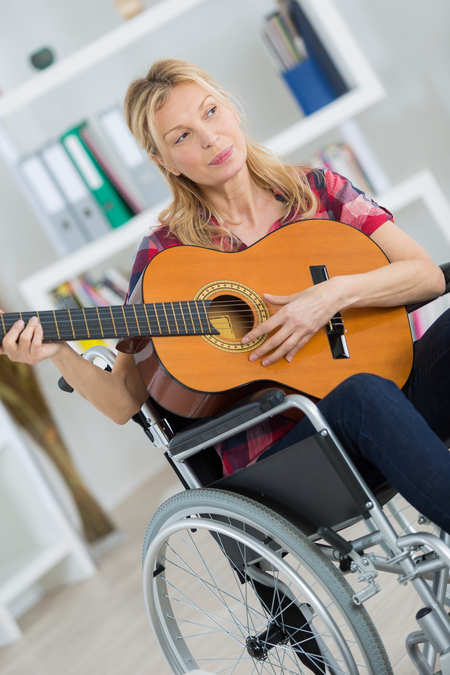 Kvinna i rullstol spelar akustisk gitarr i ett ljust rum med hyllor i bakgrunden.