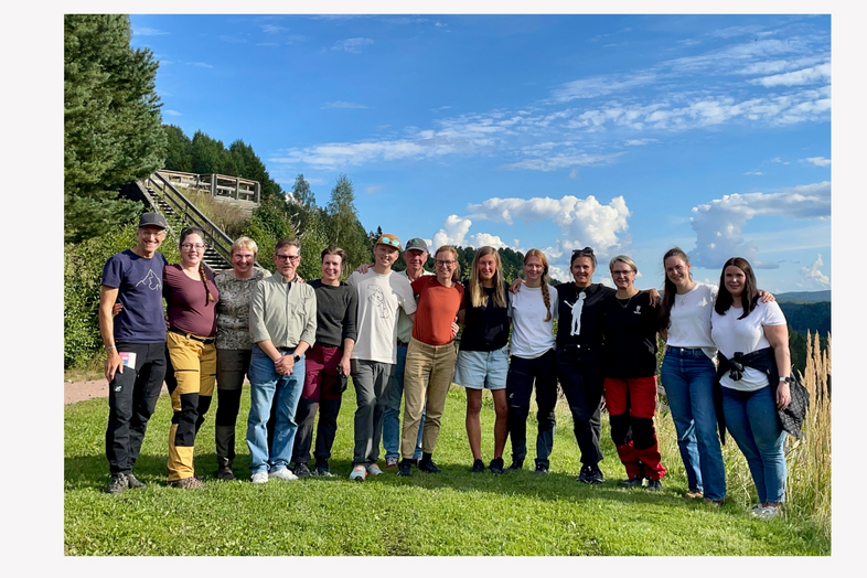 En grupp människor står tillsammans utomhus på en gräsmatta med blå himmel och träd i bakgrunden.