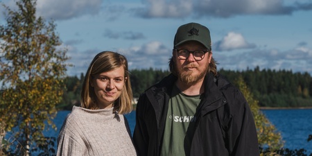 En kvinna och en man står utomhus vid en sjö med skog i bakgrunden under en solig dag.