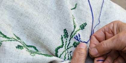 Närbild av händer som broderar med blå och gröna trådar på vitt tyg.