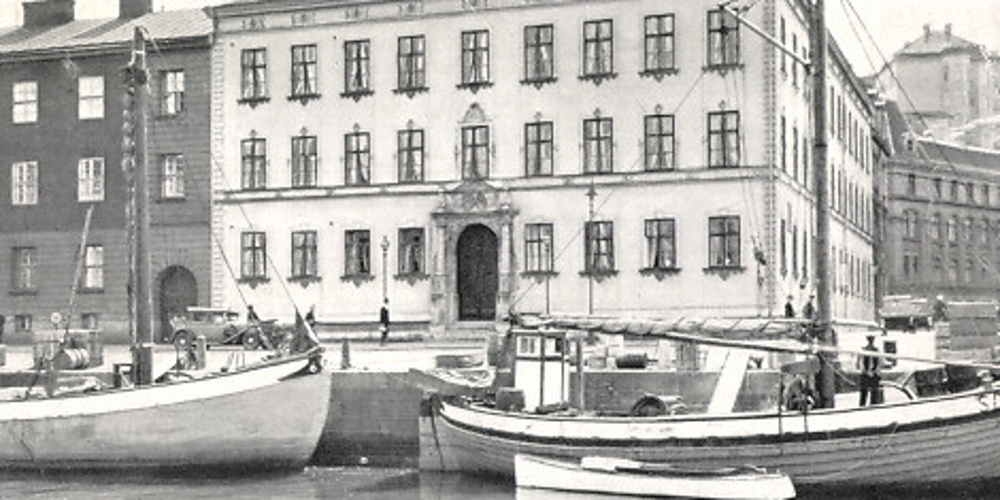 Svartvit bild av båtar vid kajen framför en stor, gammal byggnad i stadsmiljö.