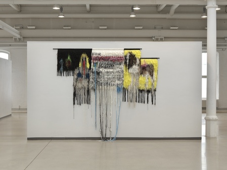 Textil konstinstallation med färgglada trådar som hänger från en vägg i ett galleri.
