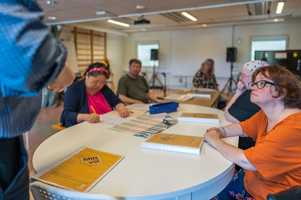 Personer sitter runt ett bord i ett klassrum och arbetar med papper och böcker.