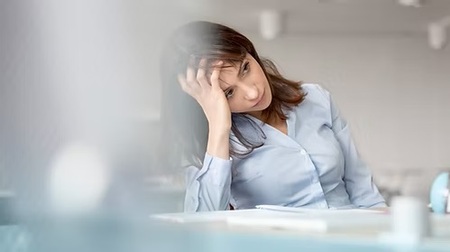 En kvinna i blå skjorta sitter vid ett skrivbord och lutar huvudet mot handen, ser trött ut.
