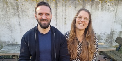 Två personer sitter på en bänk utomhus med en svart ledarhund mellan sig.