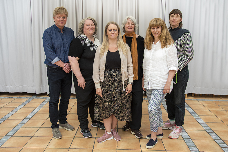 Sex vuxna personer står i en rad inomhus framför vita gardiner och tittar mot kameran.