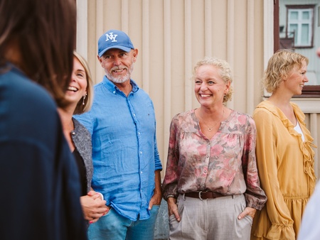 En grupp vuxna står utomhus framför en beige vägg och samtalar.