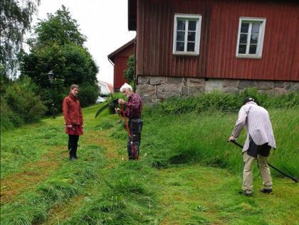 Tre personer slår gräs med liar vid ett rött trähus på landet.