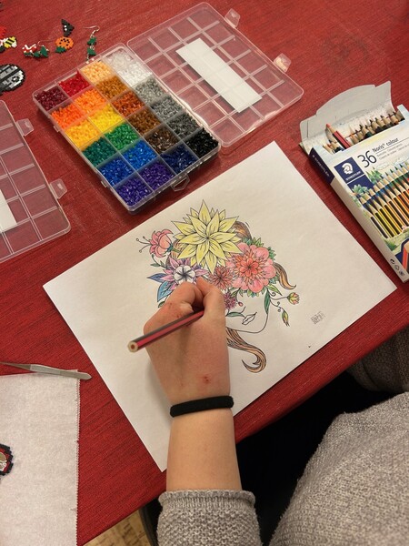 En person färglägger en teckning av ett ansikte med blommor i håret vid ett bord med färgpennor och pärlor.