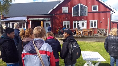 En grupp människor står utomhus framför ett rött trähus med vita fönster och veranda.