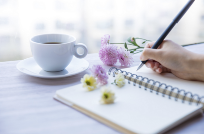 En hand skriver i en spiralbunden anteckningsbok bredvid en kopp kaffe och några blommor på ett bord.