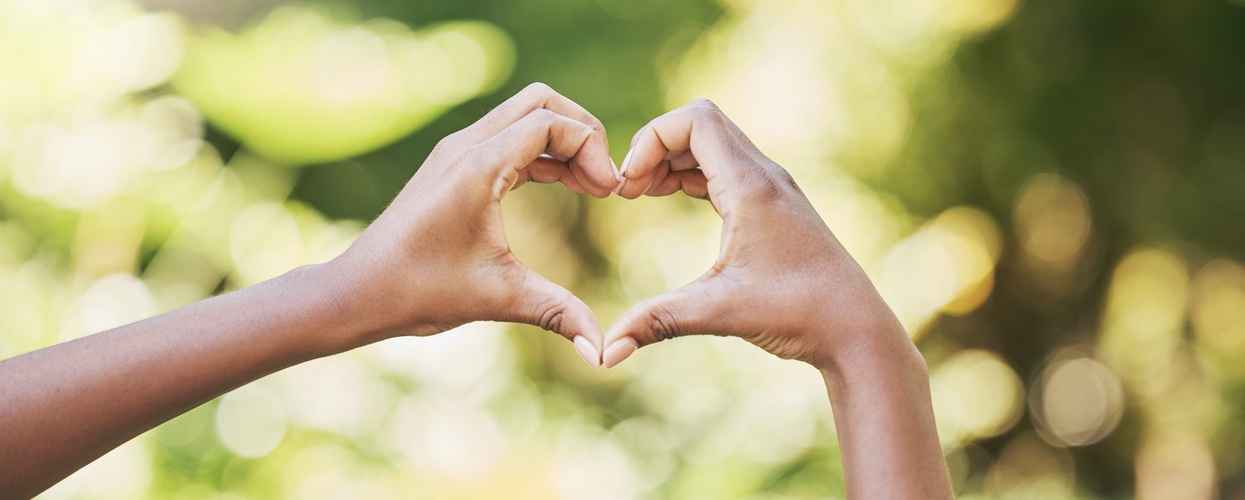 Två händer formar ett hjärta mot en suddig, grön bakgrund utomhus.