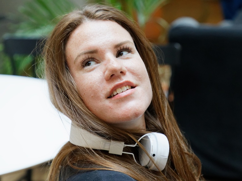 Kvinna med långt brunt hår och hörlurar runt halsen sitter inomhus och tittar uppåt.