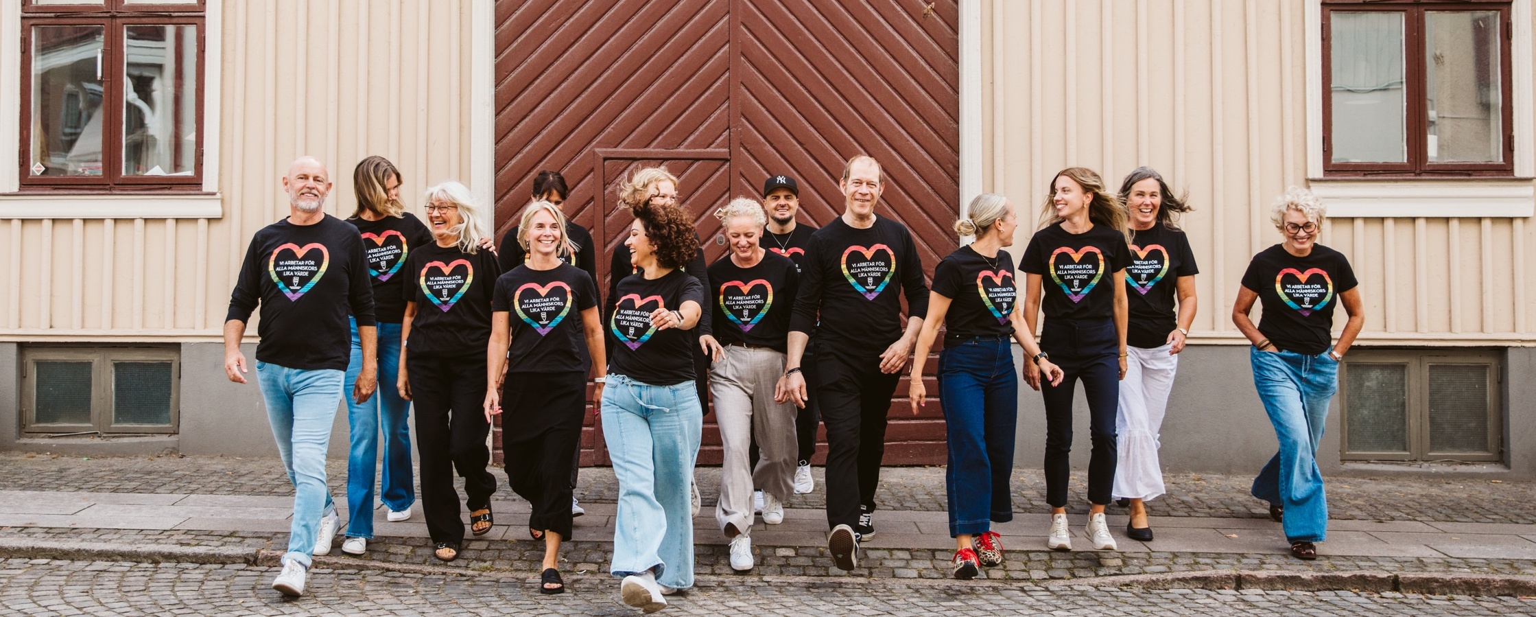 En grupp människor i svarta tröjor med regnbågshjärtan går tillsammans på en kullerstensgata framför en träbyggnad.