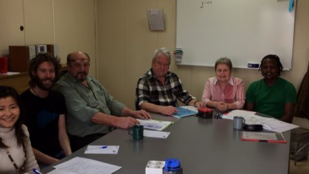 Sex personer sitter runt ett konferensbord med papper och muggar, i ett mötesrum med whiteboard.