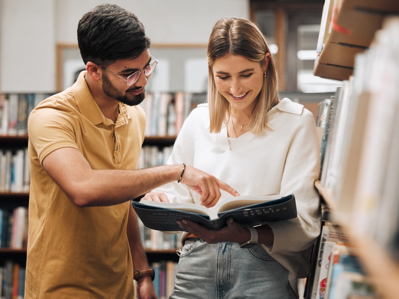 Två unga vuxna läser tillsammans i en bokhylla på ett bibliotek och ler.