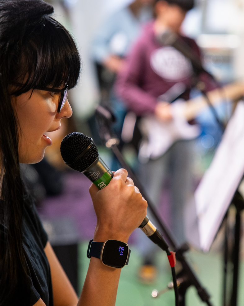 Kvinna med mörkt hår sjunger i mikrofon, person i bakgrunden spelar elgitarr.