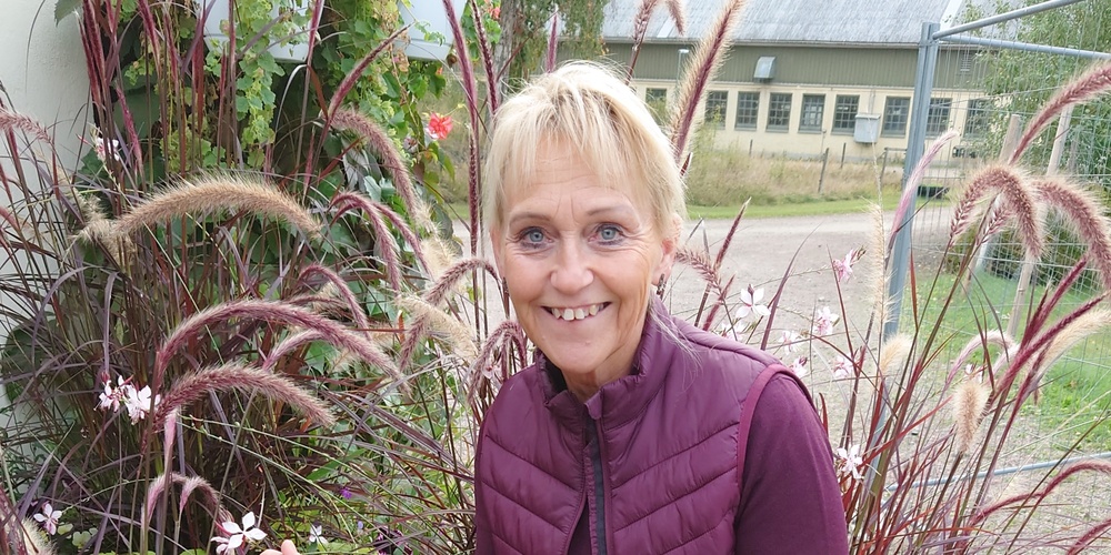 Person sitter på en blomlåda utomhus omgiven av prydnadsgräs och blommor.
