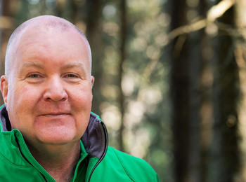 Person i grön jacka står i en solbelyst skog med träd i bakgrunden.