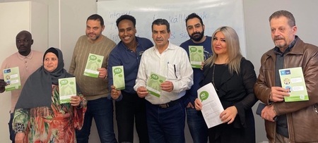 Åtta personer står i rad inomhus och håller informationsblad, framför en whiteboard.