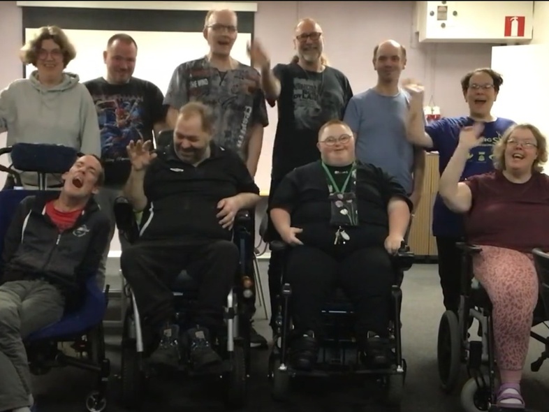 En grupp människor, några i rullstol, sitter och står tillsammans inomhus och vinkar mot kameran.