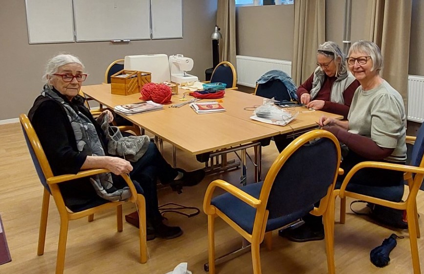 Tre personer sitter runt ett bord och handarbetar i ett ljust rum med trästolar och handarbetsmaterial.