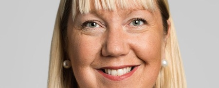 Porträtt av en leende kvinna med blont hår och lugg, mot ljus bakgrund.