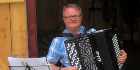 Man i glasögon spelar dragspel framför en trävägg, notställ syns i förgrunden.