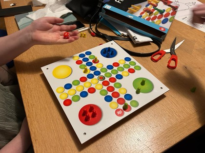 Två personer spelar brädspelet Fia med knuff vid ett träbord med spelpjäser och tärning.