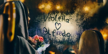 En person med huva sitter på en buss, håller blommor och tittar på ett immigt fönster där det står 'Violetta ♥ Alfredo'.
