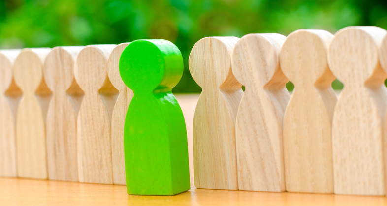 En grön träfigur står mellan två rader av ljusa träfigurer på ett bord med grön bakgrund.