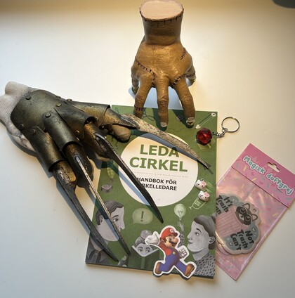 Bok med titeln 'Leda Cirkel', en konstgjord hand, Freddy Krueger-handske, Mario-sticker och nyckelring på ett bord.