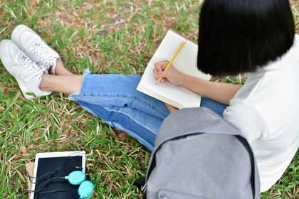Person sitter på gräs, skriver i anteckningsbok med penna. Ryggsäck, hörlurar och surfplatta bredvid.