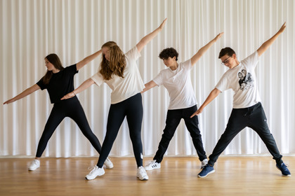 Fyra ungdomar står i rad och gör en dansrörelse med armarna utsträckta i ett ljust rum.