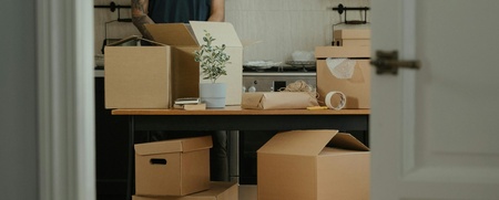 En person packar upp flyttkartonger i ett kök, sedd genom en öppen dörr.