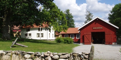 Gårdsmiljö med röd lada, vit byggnad, stengärdsgård och grönskande träd under blå himmel.