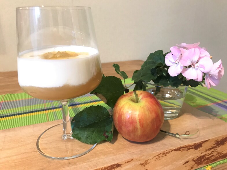 Ett glas med skummande dryck, ett äpple med blad och en rosa blomma i vas på ett träbord.