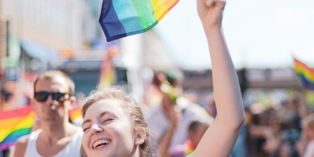 Leende person håller en regnbågsflagga på en prideparad under en solig dag.